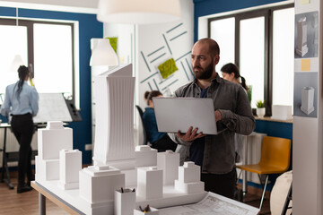 Poster - Urban architect adult inspecting design plan at workplace for professional layout of building model maquette. Engineering contractor working on laptop computer for construction