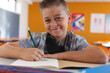 © WavebreakMediaMicro - Portrait of smiling caucasian schoolboy with face mask sitting in classroom, writing in schoolbook