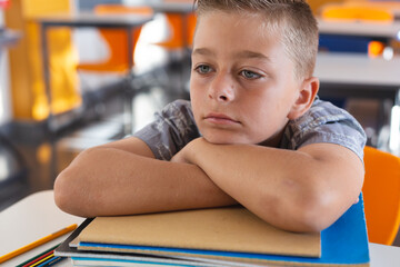 Wall Mural - Tired looking caucasian schoolboy sitting at desk in classroom leaning on a pile of schoolbooks