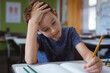 © WavebreakMediaMicro - Tired caucasian schoolboy in classroom sitting at desk, holding head and writing