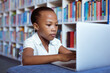 African american schoolgirl at desk in school library concentrating using laptop