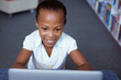 Smiling african american schoolgirl at desk in school library using laptop