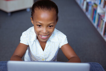 Wall Mural - Smiling african american schoolgirl at desk in school library using laptop