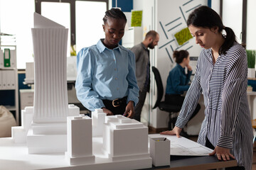 Poster - Multi ethnic women working as professional architects partners for designing blueprint plans. Colleagues standing at building model maquette urban industry design on desk.