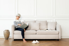 Senior Woman Reading A Book On The Sofa In A Scandinavian Decor Living Room