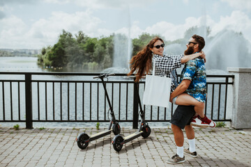 Portrait of a modern young couple on vacation having fun riding on electric scooters through the city holding white textile eco bag .