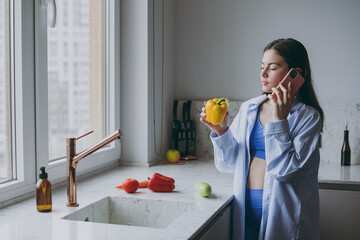 Wall Mural - Young side view pensive vegeterian housewife woman 20s in casual clothes blue shirt hold bell pepper talk mobile cell phone cooking food in light kitchen at home alone Healthy diet lifestyle concept