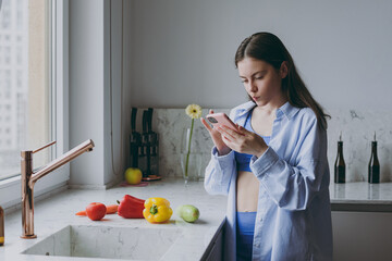 Young vegan housewife woman 20s in casual clothes blue shirt use mobile cell phone look for vegeterian recipe on internet cooking food in light kitchen at home alone Healthy diet lifestyle concept