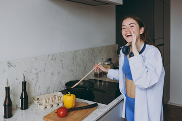 Wall Mural - Young vegeterian housewife mother woman 20s wearing casual clothes blue shirt headphones cook fried vegetables on pan calls to eat in light kitchen at home alone Healthy food diet lifestyle concept.