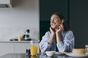 Wall Mural - Young smiling happy fun housewife woman 20s wearing casual clothes blue shirt air pods eat breakfast pancakes listen to music cook food in light kitchen at home alone Healthy diet lifestyle concept.