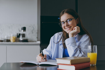 Wall Mural - Smart smiling happy young housewife woman 20s in blue shirt casual clothes glasses writing down recipe in notebook diary prop up chin cook food in light kitchen at home Healthy diet lifestyle concept