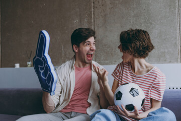 Wall Mural - Overjoyed couple friends woman man football fans 20s in casual clothes foam glove finger up support favorite team scream with soccer ball scream indoors at home watch tv People sport family concept.