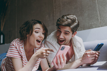 Surprised excited fun young couple two friends woman man in casual clothes lying sofa point finger on mobile cell phone surfing internet rest indoors at home flat together People lifestyle concept.