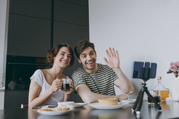 Young couple two woman man in casual clothes sit by table eat pancake talk video call mobile cell phone drink waving hand drink coffee cook food in light kitchen at home together. Healthy diet concept