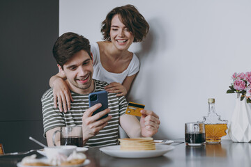 Young cheerful happy couple two woman man in casual clothes sit by table eat pancake use mobile cell phone credit bank card shop online order delivery in light kitchen at home Healthy diet concept