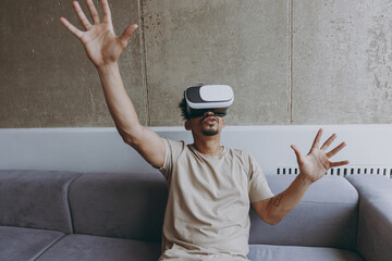 Wall Mural - Happy amazed impressed fun young african american man in beige t-shirt sit on grey sofa indoors apartment watch in vr headset pc gadget play game with outstretched hands rest on quarantine stay home.