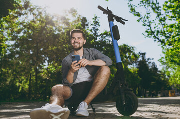 Full length bottom view young happy man in blue shirt sit near riding scooter hold mobile cell phone on alley rest in spring green city sunshine park outdoor on nature Urban leisure lifestyle concept