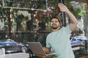 Smiling freelancer friendly happy student young man 20s in casual mint t-shirt work online with laptop pc computer use free wifi waving hand near outdoors cafe Urban leisure lifestyle lunch concept