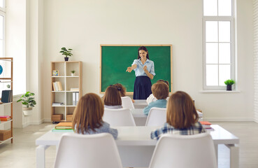 friendly smiley school teacher asks her elementary students questions in class. little children sitt