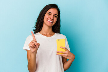 Young caucasian woman holding a mobile phone isolated on blue background showing number one with finger.