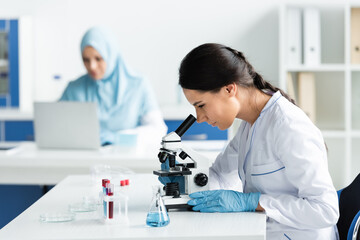 Wall Mural - Scientist in latex gloves looking through microscope near flask with reagent and test tubes