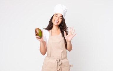 Wall Mural - pretty woman smiling and looking friendly, showing number five wearing an apron and holding a mango