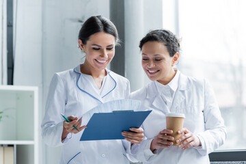 Wall Mural - Multiethnic doctors with coffee and clipboard smiling in hospital