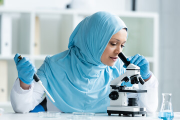 Wall Mural - Arabian scientist in hijab looking through microscope and holding electronic pipette