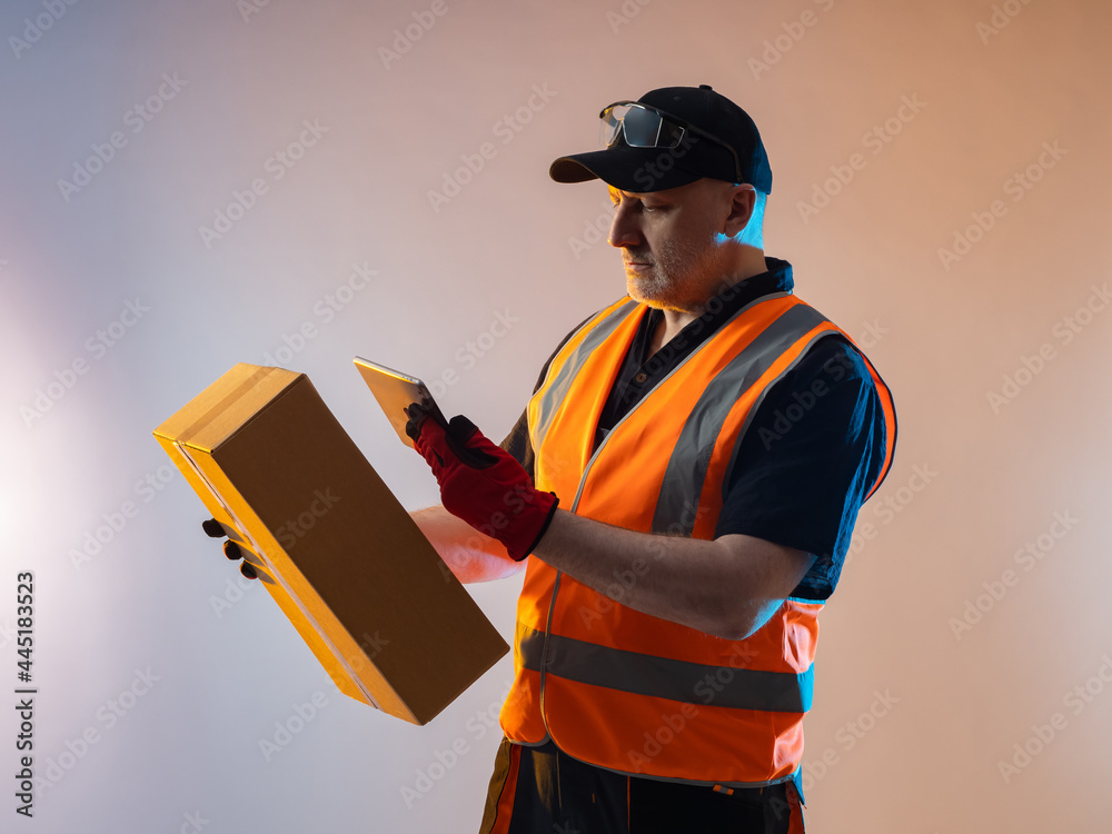 Order fulfillment process concept. A man works in an order fulfillment center. He photographs a cardboard box with his phone. An employee of an order fulfillment center in an orange vest. - obrazy, fototapety, plakaty 