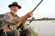 Positive gray-haired man fishing on lake in nature, patience and recreation concept.Portrait of senior caucasian fisherman with rod sitting on chair in nature, countryside, smiling at camera