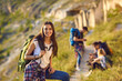 © Studio Romantic - Young smiling woman tourist hiker with backpack standing and looking at camera with group of friends hikers resting at background on nature on sunny clear day, selective focus. Hiking concept