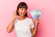 © Asier - Young mixed race woman holding bills isolated on pink background trying to listening a gossip.