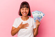 © Asier - Young mixed race woman holding bills isolated on pink background laughs out loudly keeping hand on chest.