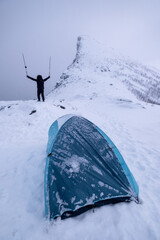 Wall Mural - mountaineer man standing and raising trekking poles on snowy hill with tent camping on the peak in b