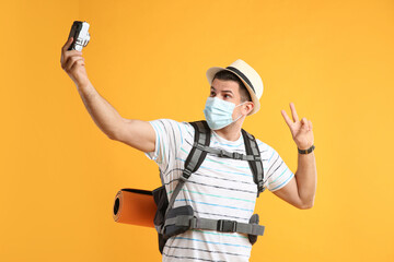 Wall Mural - Male tourist in protective mask with travel backpack taking selfie on yellow background