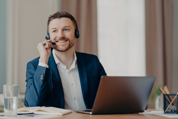 Photo of happy male manager wears headset works on laptop computer consults clients looks with glad thoughtful expression away dressed formally poses at desktop. Support and online consultation