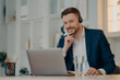 © VK Studio - Professional male operator works in callcenter uses headset and laptop computer talks on video call or virtual webcam event wears formal clothes poses against cozy interior has online meeting