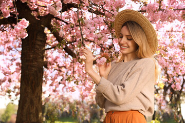 Young woman wearing stylish outfit near blossoming sakura in park. Fashionable spring look