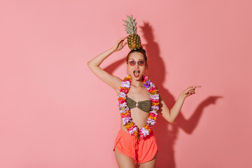 Wall Mural - Surprised lady in khaki trendy swimsuit, pink shorts and charming necklace of flowers holding on head pineapple on isolated backdrop..