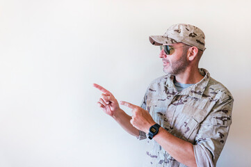 Canvas Print - Handsome man in camouflaged army uniform smiling and pointing with two hands and fingers to the side.