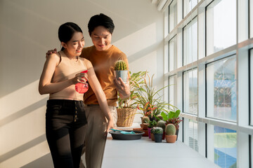 Happy Asian couples spend their free time taking care of and spraying water on the cactus plants by sunny windows in the apartment. Concept of Enjoy leisure, Favorite hobby and Indoor houseplants.