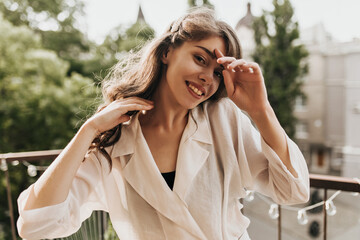 Young lady in beige outfit smiling and looking at camera on terrace. Charming pretty woman in white blouse resting on balcony