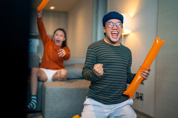 Group of Asian people friends sit on sofa watching and cheering sports games competition on TV together at home. Excited man and woman sport fans celebrate sport team victory in national sports match