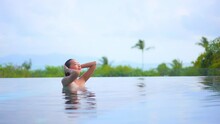 Young Pretty Exotic Woman In Infinity Swimming Pool Fixing Her Wet Hair Tropical Vegetation In Background, Full Frame Slow Motion