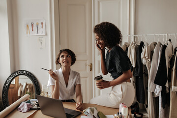 Wall Mural - Creative young fashion designers creating new clothes in cozy office. Short-haired Caucasian woman thoughtfully looks up and holds pencil. Dark-skinned lady holds coffee cup.