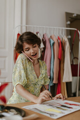 Wall Mural - Cheerful happy woman in floral v-neck dress holds pen and designs new fashion collections. Charming lady talks on phone.