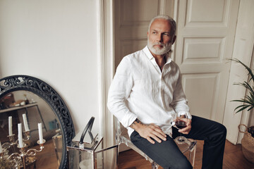 Man in white shirt sitting on chair and holding wine. Serious guy with beard in stylish outfit is posing in his apartment