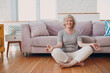 © primipil - Senior adult smiling woman practicing yoga at home living room. Elderly relaxed female sitting in lotus pose and meditating zen like