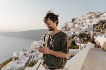 Brunette man in stylish summer outfit chats in phone outside. Guy in dark green t-shirt smiles and poses in beautiful city with sea view.