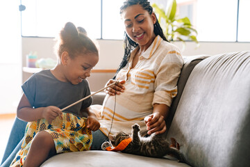 Black pregnant woman and her daughter playing with kitten on sofa